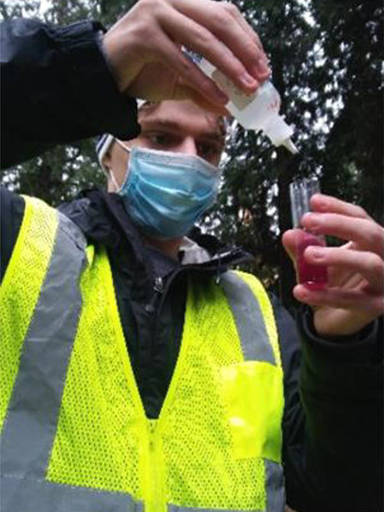 A young man with a mask and safety vest on pours testing fluid into a beaker from a squirt bottle