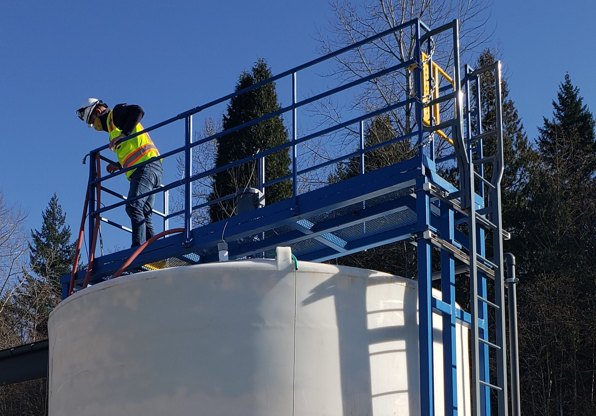 A worker in a safety vest, hardhat, and mask, inspects the magnesium hydroxide tank at the Brightwater treatment plant. The person is standing on blue scaffolding above the big, white tank
