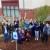 A group of 13 people smile at the camera and hold up their hands waving. They are standing or kneeling in the rain garden.